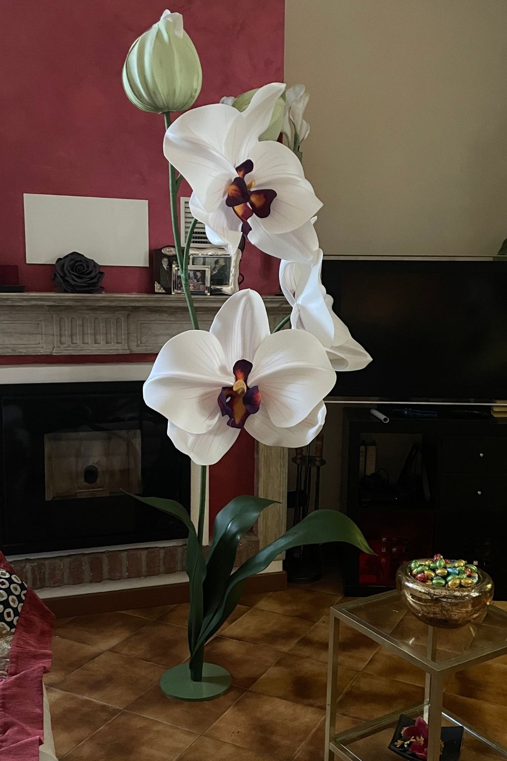 Grande scultura di orchidea bianca artigianale con cuore bordeaux e bocciolo verde, alta quasi quanto un camino in un interno domestico.