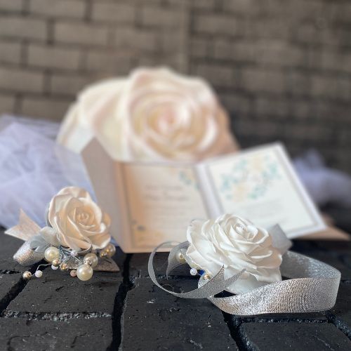 Set coordinato di accessori sposa con rose in seta avorio, perle e nastri in organza su sfondo materico scuro, stile minimal chic e sofisticato.