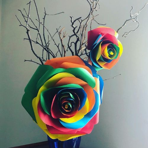 Composizione floreale con rose giganti color arcobaleno e rami secchi, scenografia artistica simbolo di inclusività e amore senza etichette.