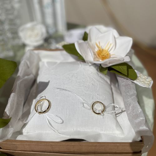 Cuscino portafedi in tessuto bianco decorato con una magnolia artigianale fatta a mano e foglie verdi, stile botanico elegante e senza tempo.