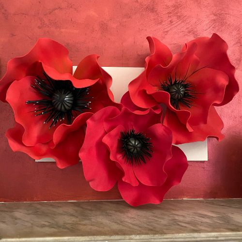 Installazione artistica a parete con tre papaveri giganti rossi e stami neri, scenografia floreale materica su fondo rosso cremisi.