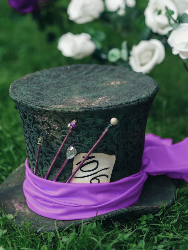 Cappello a cilindro artigianale del Cappellaio Matto in tessuto damascato verde scuro, con fascia viola, spille gioiello e cartellino "10/6", immerso in una scenografia floreale.