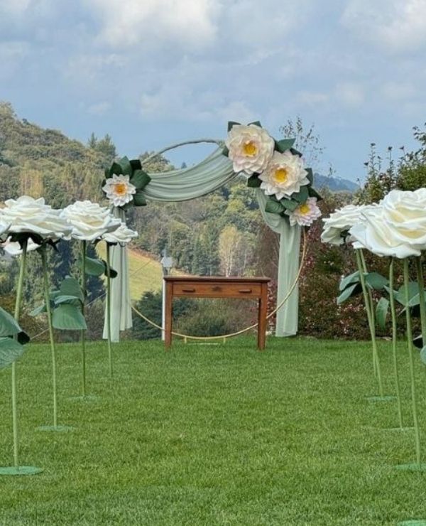 Arco nuziale circolare decorato con grandi fiori bianchi e drappeggi verde salvia, affiancato da un corridoio di rose giganti autoportanti su un prato verde.