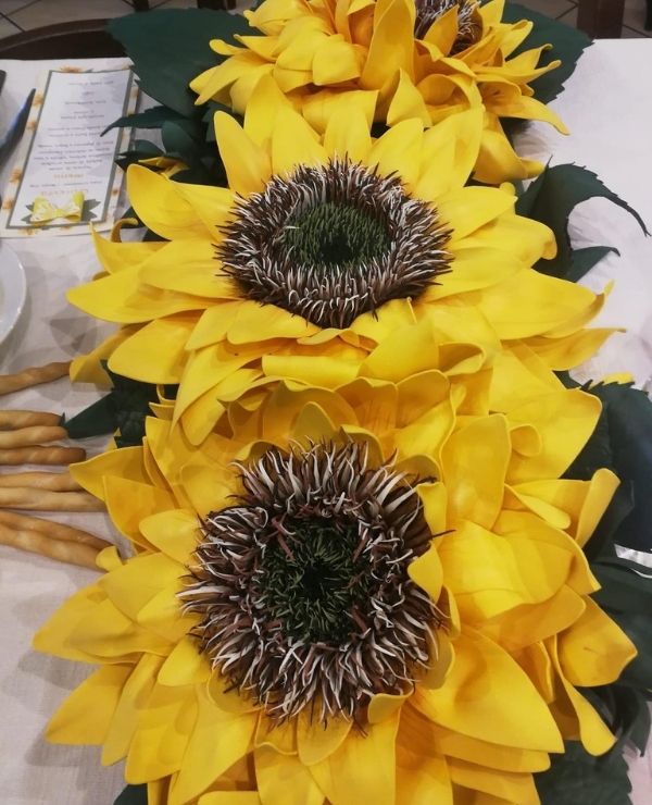 Dettaglio dall'alto di tre girasoli giganti artigianali in fommy giallo e marrone, disposti in fila come centrotavola su una tovaglia bianca.