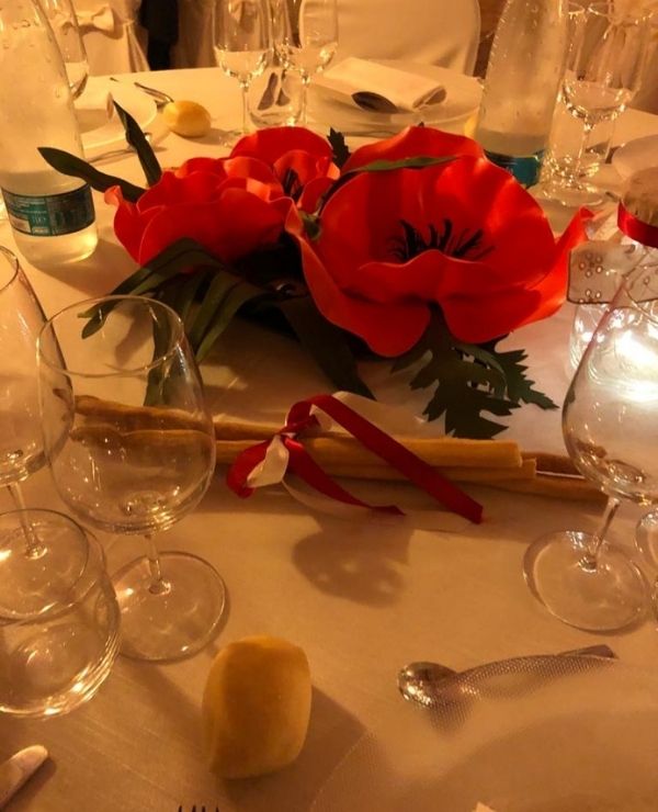 Dettaglio di un centrotavola artistico con papaveri giganti in rosso vibrante, realizzati a mano con tecnica artigianale. I fiori sovrastano la mise en place elegante di un ricevimento di nozze, creando un contrasto cromatico deciso tra il rosso fuoco dei petali e il bianco della tovaglia.