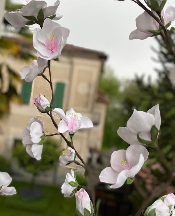 Dettaglio ravvicinato di fiori di pesco giganti fatti a mano in materiale tecnico bianco con sfumature rosa pesca, montati su rami scenografici, con sfondo sfocato di una villa storica