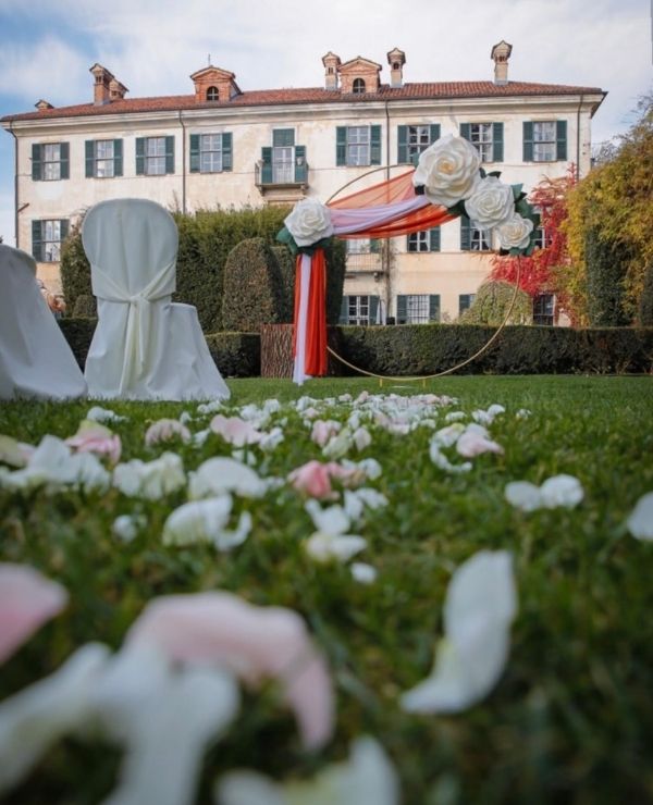 Allestimento per cerimonia all'aperto nel giardino di una villa storica con arco circolare decorato da rose bianche giganti e tessuti color ruggine, sedie bianche e petali bianchi e rosa sull'erba.