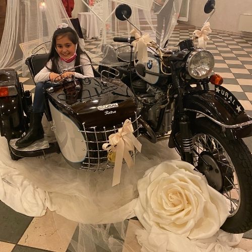 Sidecar d’epoca nero Ural decorato con fiocchi di raso color panna e una rosa gigante artigianale bianca alla base della ruota anteriore. Una bambina sorridente è seduta nel carrozzino su uno sfondo di tulle bianco e pavimento a scacchi.