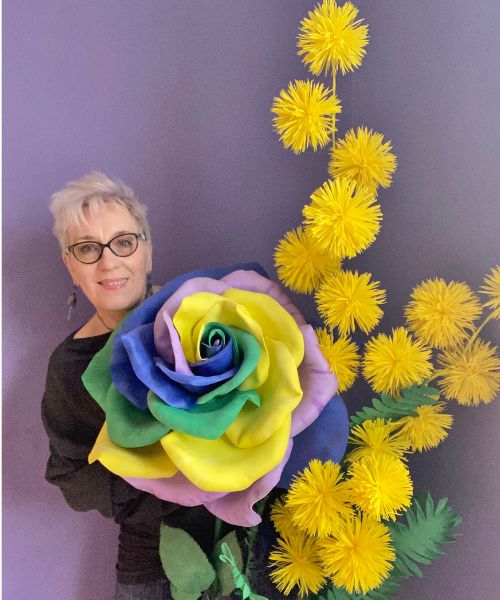 Donna con occhiali che sorregge una grande rosa scenografica con petali color arcobaleno e mimosa gigante su sfondo lilla.
