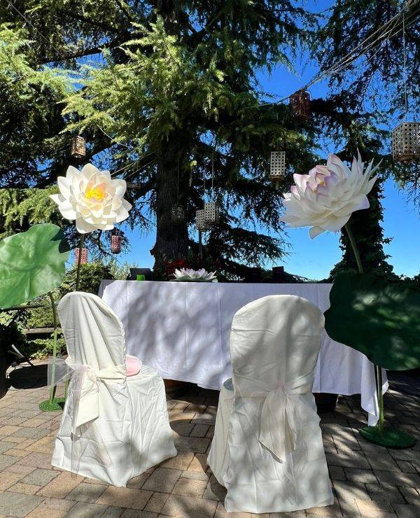 Allestimento wedding scenografico con fiori di loto giganti realizzati a mano, posizionati ai lati di un tavolo cerimoniale sotto un grande albero secolare. I fiori bianchi e sfumati sovrastano le sedute, creando un’atmosfera onirica e surreale ispirata al mondo di Alice nel Paese delle Meraviglie.